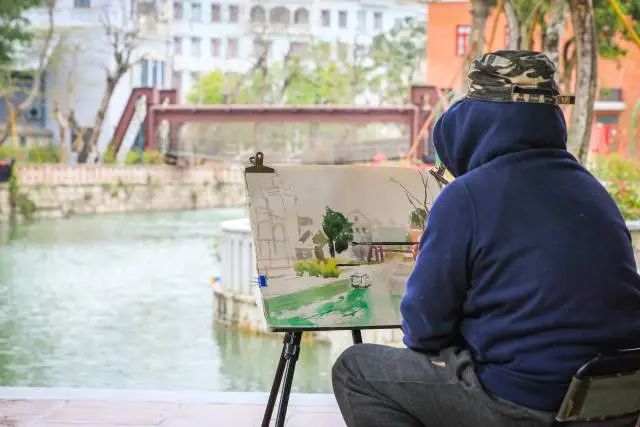 小橋流水的一側,一位民間畫家落筆畫下了古鎮的綠美景象。 圖片